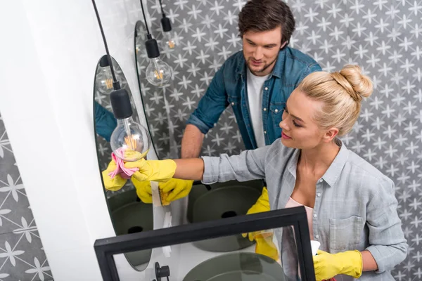 Attractive girl and handsome man in rubber gloves cleaning mirror in bathroom — Stock Photo