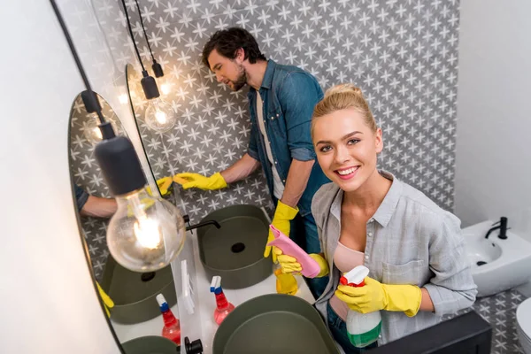Sonriente chica en guantes de goma sosteniendo spray y trapo mientras marido limpieza espejo en cuarto de baño - foto de stock