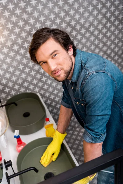 Hombre guapo en guantes de goma fregadero de limpieza y mirando a la cámara en el baño - foto de stock