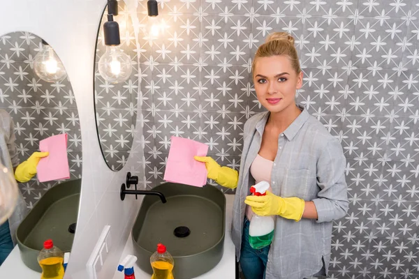 Chica sonriente en guantes de goma sosteniendo trapo y spray y mirando a la cámara en el baño - foto de stock