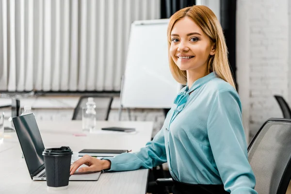 Боковой вид улыбающейся бизнесвумен на рабочем месте с ноутбуком и кофе, чтобы пойти в офис — стоковое фото