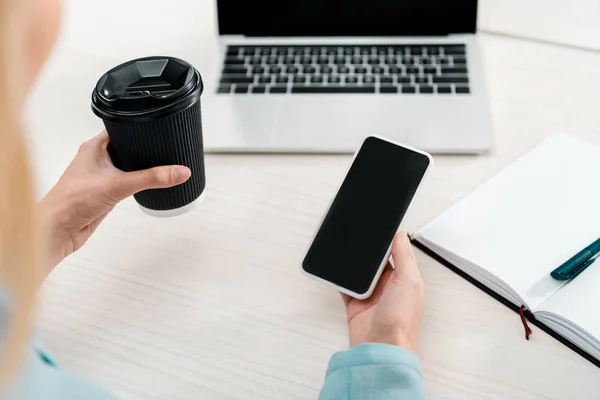 Partial view of businesswoman with coffee to go and smartphone with blank screen at workplace — Stock Photo