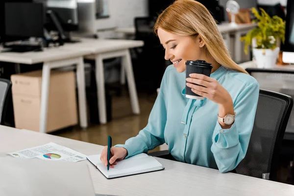 Mujer de negocios sonriente con café para ir tomando notas en el cuaderno en el lugar de trabajo en la oficina - foto de stock