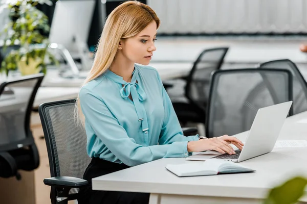 Side view of young businesswoman working on laptop at workplace with notebook in office — Stock Photo