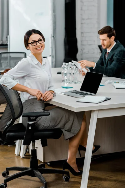 Selektiver Fokus einer lächelnden Geschäftsfrau mit Brille, die im Büro mit multiethnischen Kollegen in die Kamera blickt — Stockfoto