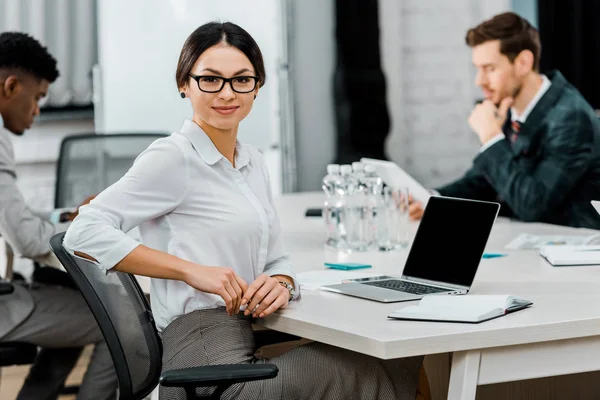 Selective focus of smiling businesswoman in eyeglasses looking at camera in office with multiracial colleagues — Stock Photo