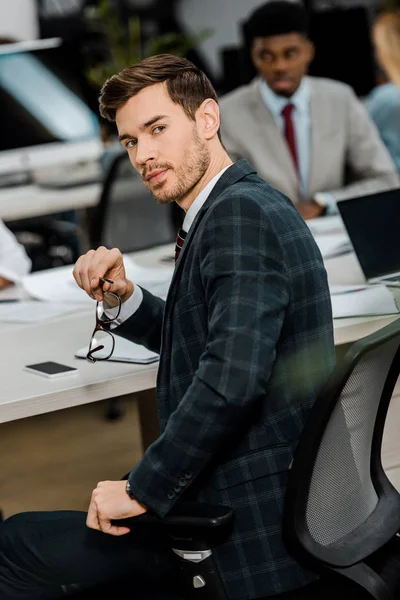 Selektiver Fokus eines jungen Geschäftsmannes im Anzug, der am Arbeitsplatz im Büro in die Kamera blickt — Stockfoto