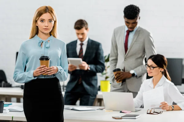 Selective focus of businesswoman with coffee to go and multiethnic colleagues in office — Stock Photo