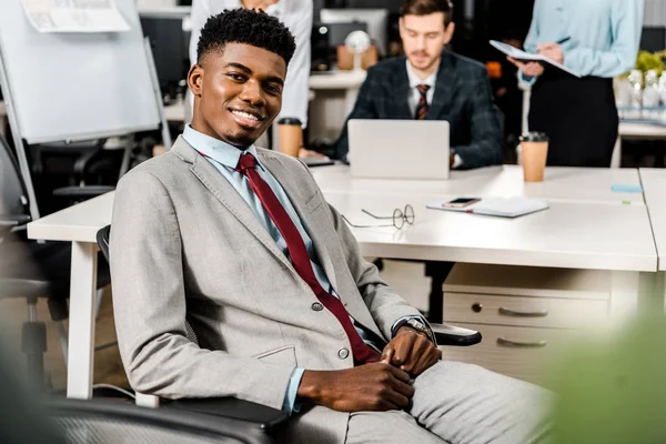 Enfoque selectivo de sonriente afroamericano empresario y colegas detrás en el lugar de trabajo en la oficina - foto de stock