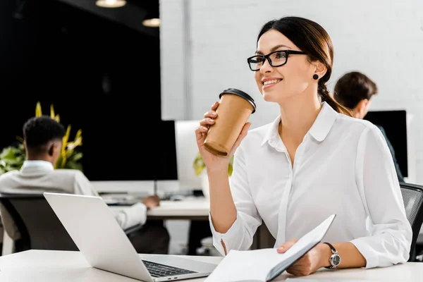 Junge Geschäftsfrau in Brille mit Coffee to go und Notizbuch am Arbeitsplatz mit Laptop im Büro — Stockfoto