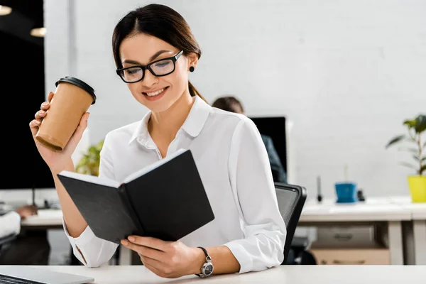 Jeune femme d'affaires en lunettes avec café à emporter et carnet de notes au travail au bureau — Photo de stock