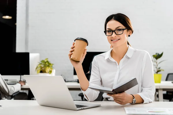 Jovem empresária em óculos com café para ir e notebook no local de trabalho com laptop no escritório — Fotografia de Stock