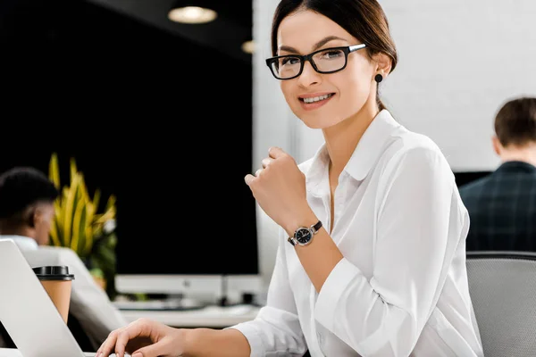 Donna d'affari sorridente in occhiali a posto di lavoro con computer portatile in ufficio — Foto stock