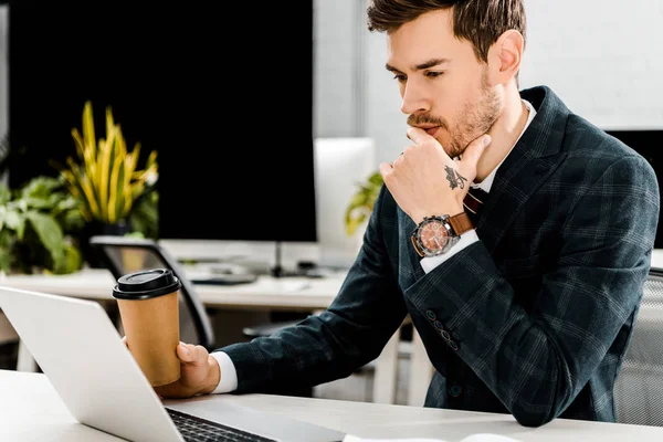 Empresário pensativo com café para ir olhando para laptop no local de trabalho em offce — Fotografia de Stock