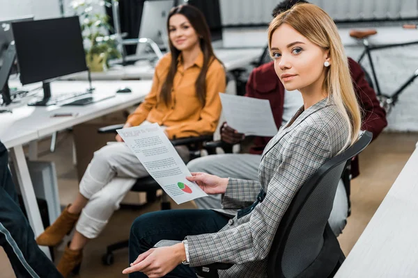 Selective focus of young businesswoman with papers near multiracial colleagues at workplace in office — Stock Photo