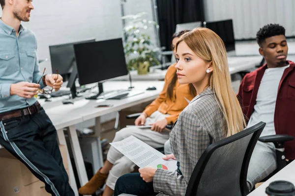 Selektiver Fokus der jungen Geschäftsfrau mit Papieren in der Nähe multiethnischer Kollegen am Arbeitsplatz im Büro — Stockfoto