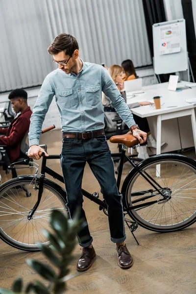 Selective focus of young businessman leaning on bicycle in office with multiracial colleagues behind — Stock Photo