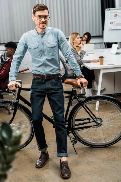 Foco seletivo do jovem empresário inclinado na bicicleta no escritório com colegas multirraciais por trás — Fotografia de Stock