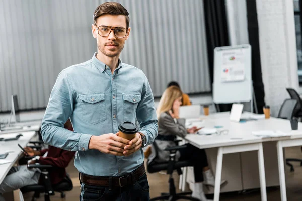 Selective focus of young businessman with coffee to go in office with multiracial colleagues behind — Stock Photo