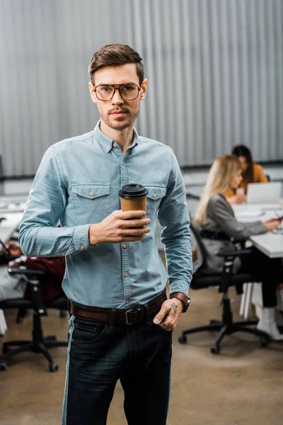 Selective focus of young businessman with coffee to go in office with multiracial colleagues behind — Stock Photo