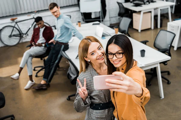 Smiling businesswomen taking selfie on smartphone in office with multiracial colleagues behind — Stock Photo