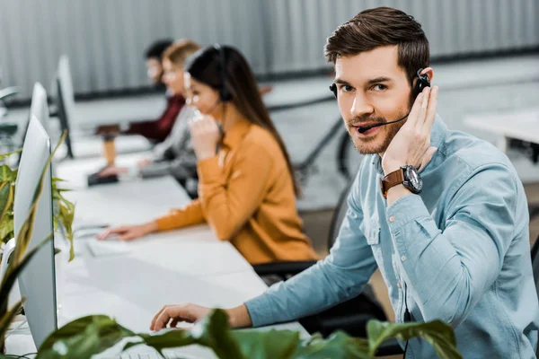 Selective focus of call center operator looking at camera while multiethnic colleagues working in office — Stock Photo