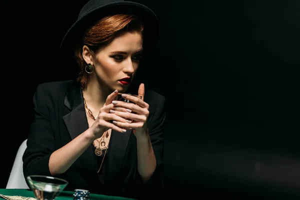 Chica atractiva en chaqueta y sombrero sosteniendo vaso de whisky en la mesa de póquer en el casino y mirando hacia otro lado - foto de stock