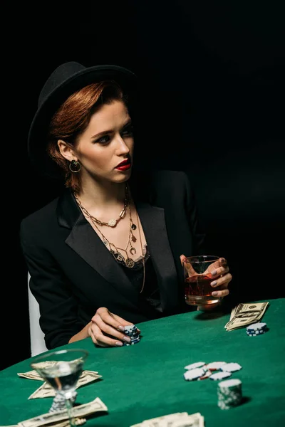 Attractive girl in jacket and hat holding glass of whiskey at poker table in casino — Stock Photo