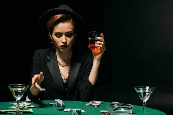 Attraktives Mädchen in Jacke und Hut, das ein Glas Whiskey in der Hand hält und im Casino Poker spielt — Stockfoto