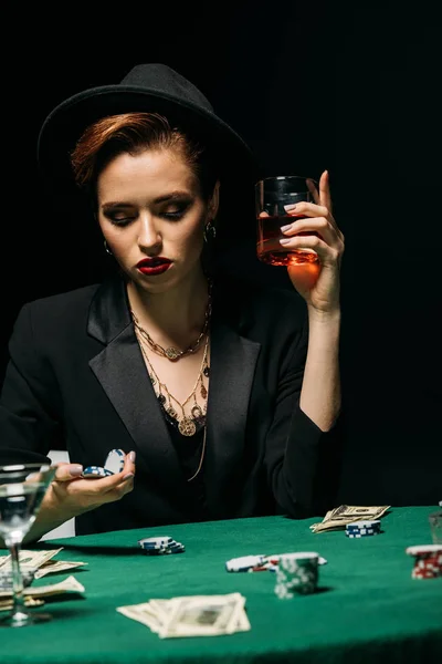 Menina bonita em casaco e chapéu segurando copo de whisky e fichas de poker à mesa no casino — Fotografia de Stock