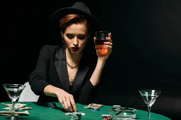 Hermosa chica en chaqueta y sombrero sosteniendo vaso de whisky y jugando al póquer en el casino - foto de stock