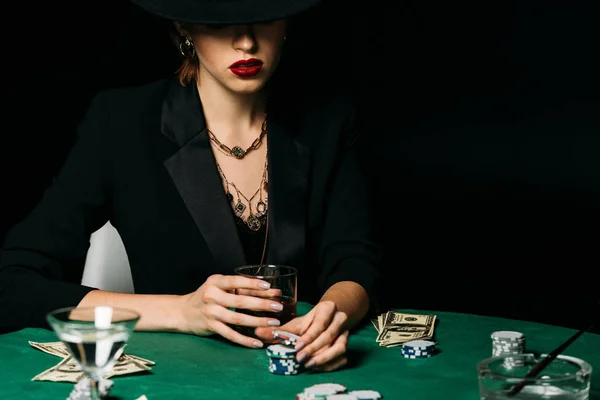 Chica atractiva en chaqueta y sombrero sosteniendo vaso de whisky y tomando ficha de póquer en el casino - foto de stock