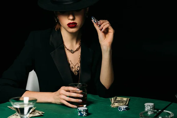 Привлекательная девушка в куртке и шляпе держа стакан виски и фишки для покера за столом в казино — стоковое фото