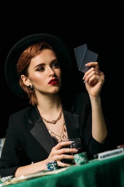 Nivel de superficie de la hermosa chica en chaqueta y sombrero sosteniendo vaso de whisky y jugar al póquer en el casino - foto de stock