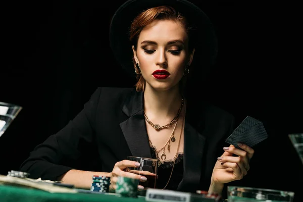 Nível de superfície da menina atraente na jaqueta e chapéu segurando copo de uísque e jogar poker no cassino — Fotografia de Stock