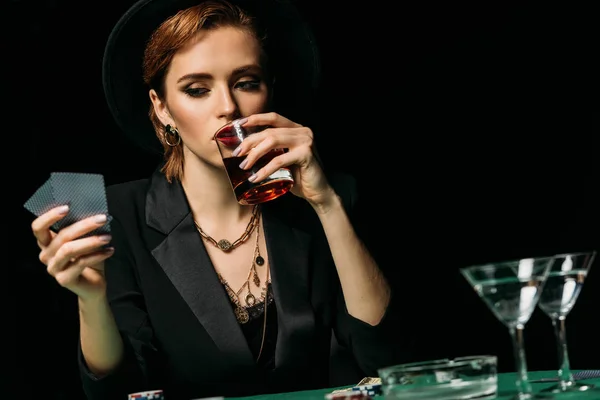 Chica atractiva en chaqueta y sombrero bebiendo whisky y mirando las tarjetas de póquer en el casino - foto de stock