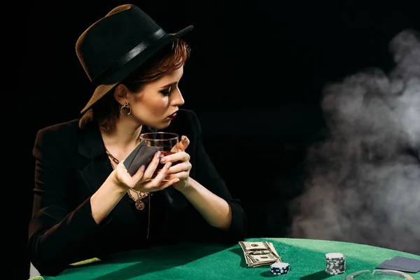 Jolie fille en veste et chapeau tenant verre de whisky tout en jouant au poker à table dans le casino, détournant les yeux — Photo de stock