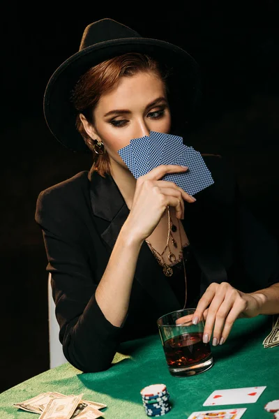 Attraktives Mädchen in Jacke und Hut, das Gesicht mit Pokerkarten im Casino verdeckt — Stockfoto