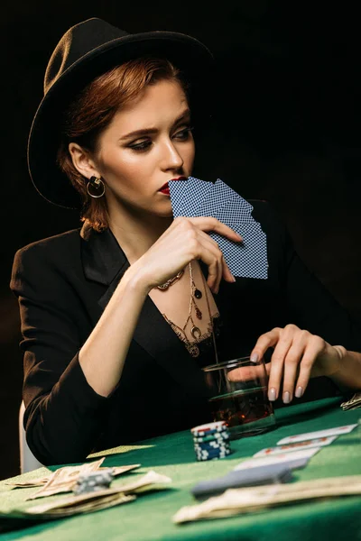 Nivel de superficie de chica atractiva en chaqueta y sombrero cubriendo la cara con tarjetas de póquer en el casino - foto de stock