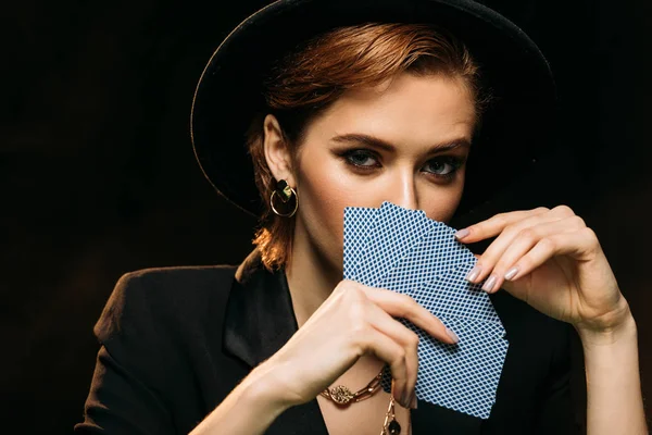 Attraktives Mädchen in Jacke und Hut, das Gesicht mit Pokerkarten isoliert auf schwarz verdeckt, blickt in die Kamera — Stockfoto