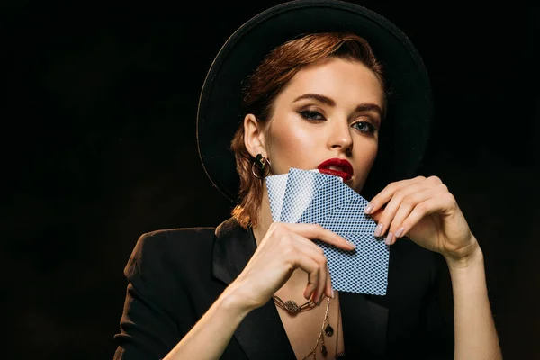 Beautiful girl in jacket and hat covering face with poker cards and looking at camera isolated on black — Stock Photo