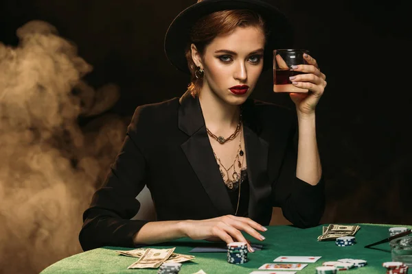 Attractive girl in jacket and hat holding glass of whiskey at poker table in casino and looking at camera — Stock Photo