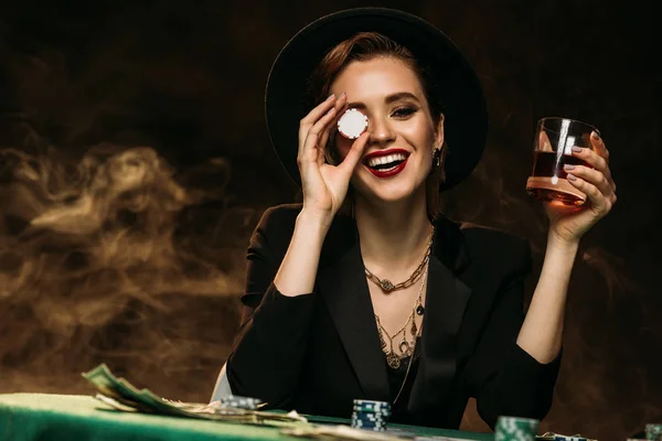 Chica atractiva feliz en chaqueta y sombrero sosteniendo vaso de whisky en la mesa de póquer y ojo de cubierta con chip de casino - foto de stock