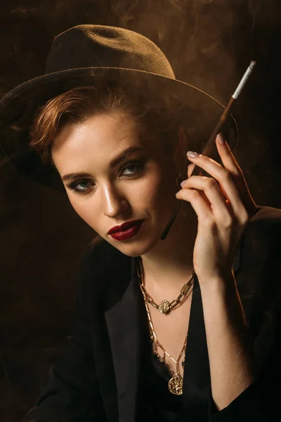 Chica atractiva en chaqueta y sombrero fumar cigarrillo en negro y mirando a la cámara - foto de stock