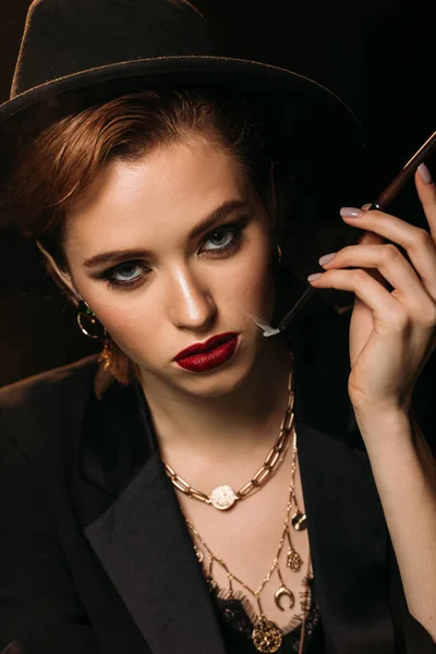 Portrait of attractive girl in jacket and hat smoking cigarette on black and looking at camera — Stock Photo