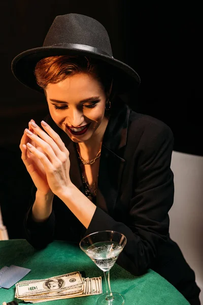 Приваблива усміхнена дівчина в капелюсі сидить за столом покеру в казино, доларові банкноти і коктейль на столі — стокове фото