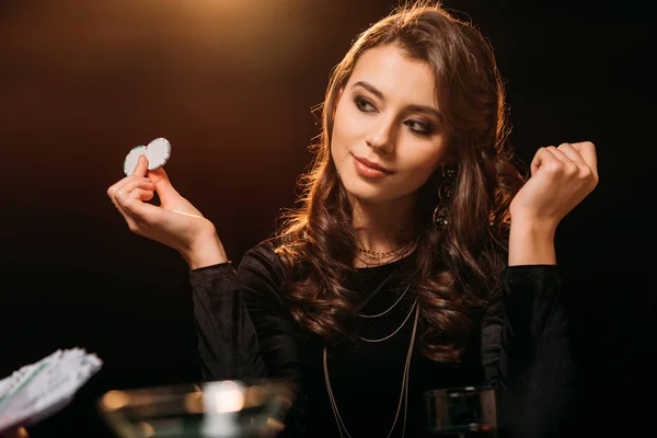 Chica atractiva sosteniendo fichas de póquer y mirando a la mesa de póquer en el casino - foto de stock