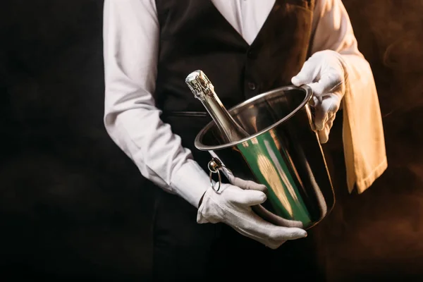 Immagine ritagliata di cameriere in possesso di bottiglia di champagne alcol in secchio su nero — Foto stock