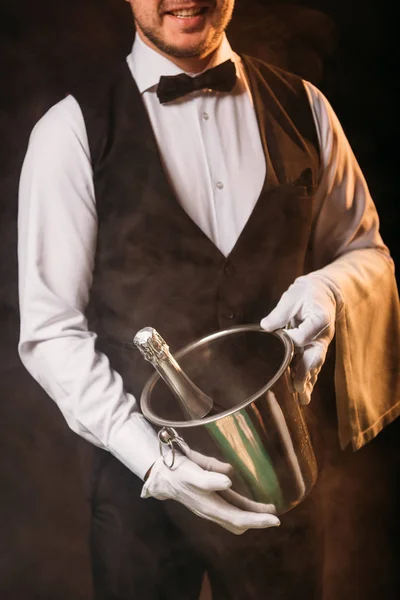 Cropped image of smiling waiter holding bottle of champagne in bucket on black — Stock Photo