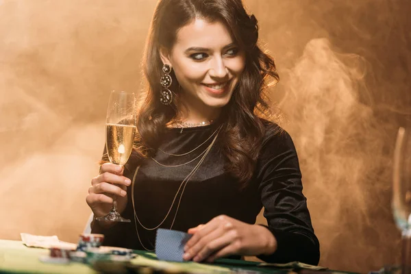 Atractiva chica sonriente sosteniendo una copa de champán en la mesa de póquer en el casino y mirando hacia otro lado - foto de stock